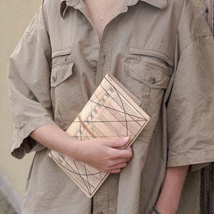 Pode incluir: Uma pessoa com cabelo castanho encaracolado vestindo uma camisa marrom clara e segurando um diário de couro marrom com um design geométrico.