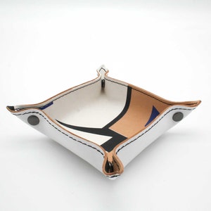 May include: A white leather tray with a geometric pattern in brown, black, and blue. The tray has a stitched edge and four metal rivets.