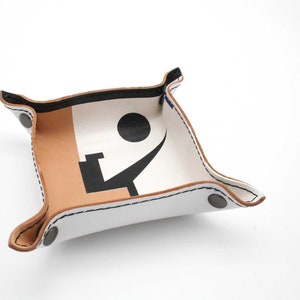 May include: A white leather tray with a brown leather trim. The tray has a black and white design on the inside. The design includes a circle and a curved line. The tray has two black metal rivets on the side.