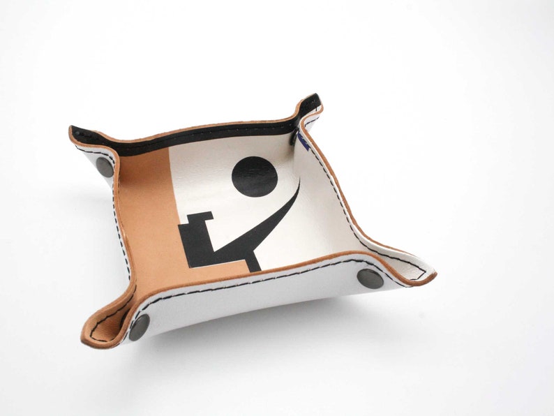 May include: A white leather tray with a brown leather trim. The tray has a black and white design on the inside. The design includes a circle and a curved line. The tray has two black metal rivets on the side.