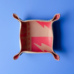 May include: A small, square, leather tray with a red and yellow pattern. The tray has four flaps that fold up to create a small, square box. The tray is on a blue background.