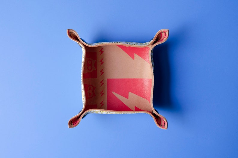 May include: A small, square, leather tray with a red and yellow pattern. The tray has four flaps that fold up to create a small, square box. The tray is on a blue background.