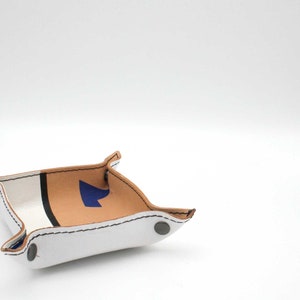 May include: A white leather tray with a brown leather accent and a blue arrow design. The tray has a white stitched border and silver metal rivets.