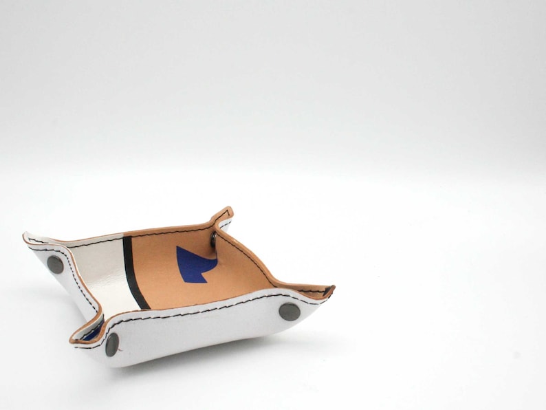 May include: A white leather tray with a brown leather accent and a blue arrow design. The tray has a white stitched border and silver metal rivets.
