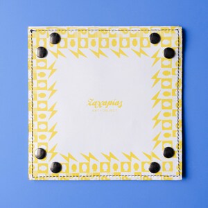 May include: A white leather tray with a yellow geometric pattern border. The tray has black metal rivets around the edge. The text 'Zaxapnias Art + Jewelry' is printed in the center of the tray.