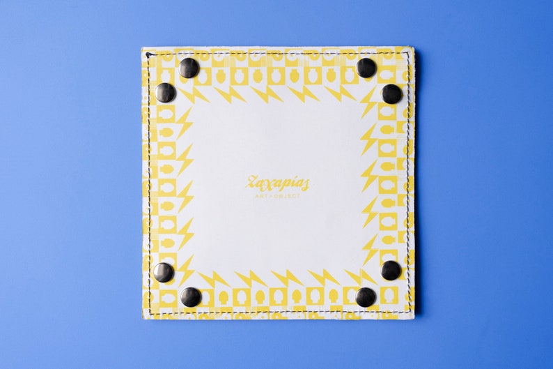 May include: A white leather tray with a yellow geometric pattern border. The tray has black metal rivets around the edge. The text 'Zaxapnias Art + Jewelry' is printed in the center of the tray.