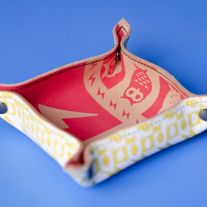 May include: A small, square, white leather tray with yellow and red geometric patterns. The tray has a red interior and two silver snaps on the sides.