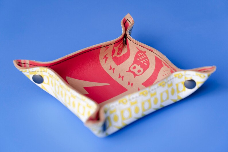 May include: A small, square, white leather tray with yellow and red geometric patterns. The tray has a red interior and two silver snaps on the sides.