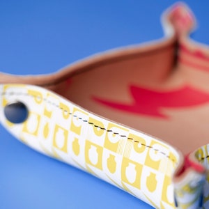 May include: A white leather tray with a yellow and white geometric pattern. The tray has a red lightning bolt design on the inside and black stitching around the edges. The tray is on a blue background.