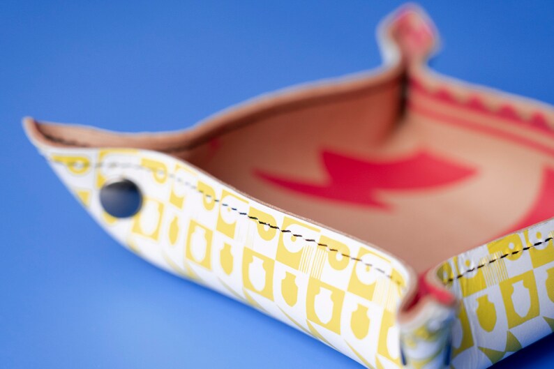 May include: A white leather tray with a yellow and white geometric pattern. The tray has a red lightning bolt design on the inside and black stitching around the edges. The tray is on a blue background.