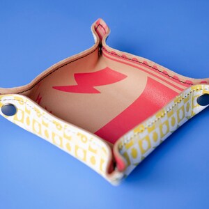 May include: A small, square-shaped leather tray with a yellow and white geometric pattern. The tray has a red lightning bolt design on the inside. The tray has a folded edge and is made of leather with a stitched seam.