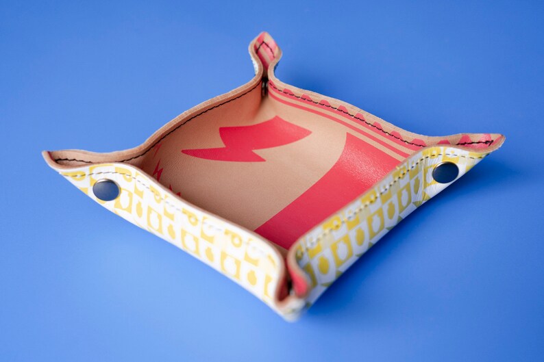 May include: A small, square-shaped leather tray with a yellow and white geometric pattern. The tray has a red lightning bolt design on the inside. The tray has a folded edge and is made of leather with a stitched seam.