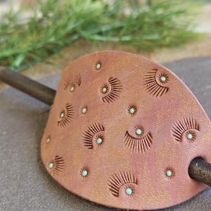 May include: A tan leather hair slide featuring a repeating pattern of stamped sunburst motifs and small, inlaid turquoise-colored dots. A dark wooden stick is inserted through the leather piece to secure it in place.