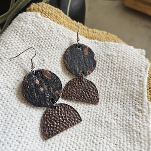 May include: Handmade earrings featuring a circular black and copper-toned top piece and a textured, copper-colored half-circle pendant. The earrings hang from silver-colored fish hook ear wires. The earrings are displayed on a white, textured surface.
