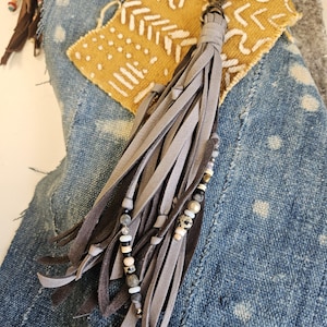 May include: A gray leather tassel keychain with a beaded accent. The tassel is made of long, thin strips of gray leather and is attached to a metal clasp. The beaded accent features a mix of white, black, and gray beads. The background is a denim fabric.