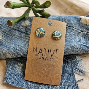 May include: A pair of round stud earrings featuring a marbled pattern in shades of teal, white, and tan, set in bronze-toned metal. The earrings are attached to a rectangular brown cardboard display card printed with the text NATIVE LEATHER CO.