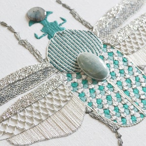 May include: A close-up of a green and silver embroidered beetle with a turquoise gemstone on its back. The beetle's wings are made of silver sequins and beads.