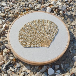 Op de afbeelding: Een witte borduurring met een gouden en witte parelmoer schelp ontwerp. De ring ligt op een achtergrond van kleine witte en grijze stenen.