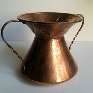 May include: A hammered copper vase with two twisted wire handles. The vase has a wide, flared top and a narrow base.