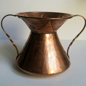 May include: A hammered copper vase with two twisted wire handles. The vase has a wide mouth and a narrow base.