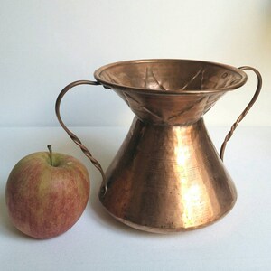 May include: A hammered copper vase with two twisted handles. The vase has a wide, flared top and a narrow base. The vase is a light copper color and has a rustic, antique look.