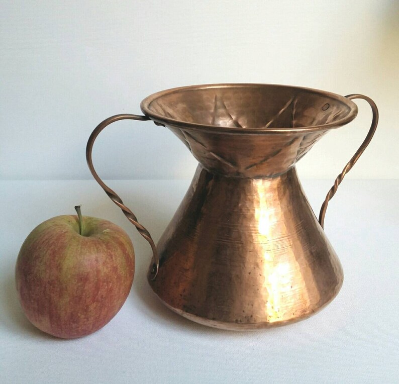May include: A hammered copper vase with two twisted handles. The vase has a wide, flared top and a narrow base. The vase is a light copper color and has a rustic, antique look.