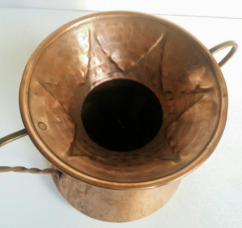 May include: A hammered copper planter with two twisted wire handles. The planter has a wide, flared opening and a rounded bottom.