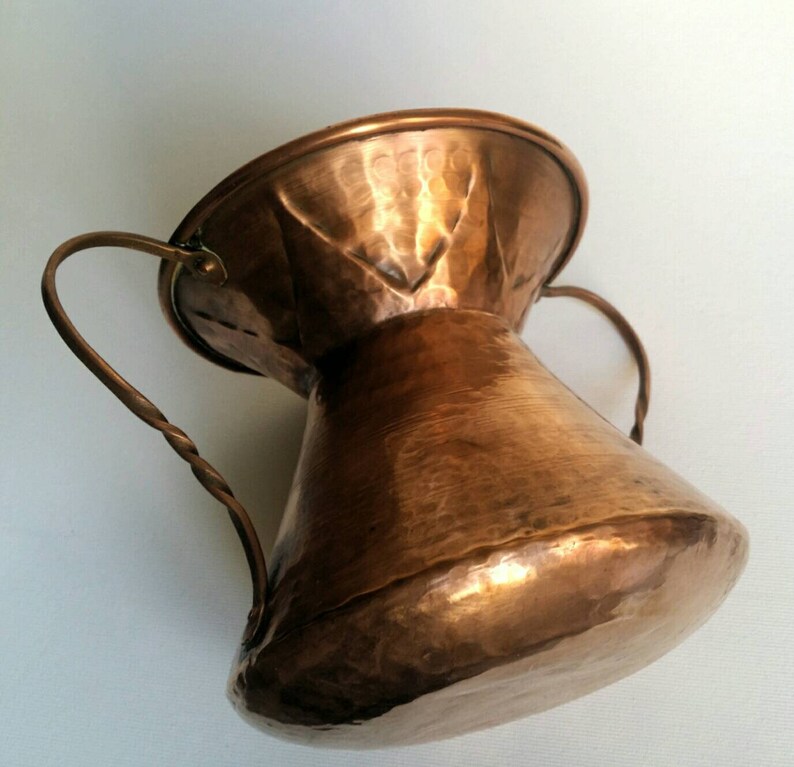 May include: A hammered copper pitcher with two twisted wire handles. The pitcher has a wide mouth and a narrow base.