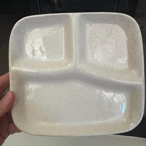May include: A square ceramic divided plate with rounded corners, featuring a glossy off-white finish with small dark speckles throughout. The plate is partitioned into three distinct sections, with two smaller compartments at the top and one larger compartment at the bottom.