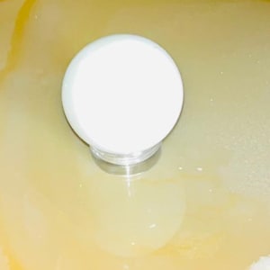 May include: A white, round object with a silver base is centered in a light yellow liquid. The object appears to be a container or a lid. The liquid has a reflective surface.