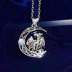 May include: Silver-toned pendant necklace featuring a crescent moon design with a dragon silhouette. The pendant has a small, round, opalescent stone and hangs from a delicate chain. The background is a dark blue.