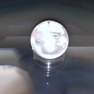 May include: A clear, round, plastic container with a lid. The container is reflecting light, creating a bright, glowing effect. The product is sitting on a reflective surface.