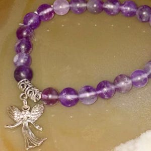 May include: A stretch bracelet composed of round, polished purple amethyst beads. The bracelet features a silver-toned metal spacer bead and a dangling silver-toned metal angel charm with outstretched wings and a long, flowing gown design.