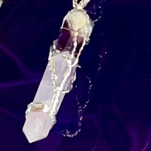 Puede incluir: Un collar con colgante de cristal con un colgante de cristal rosa claro, morado y transparente envuelto en alambre plateado. El colgante cuelga de una delicada cadena plateada, sobre un fondo morado oscuro.