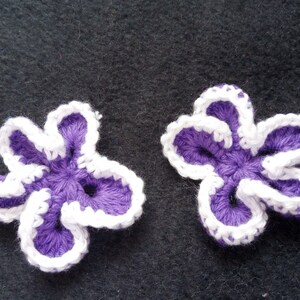 May include: Two purple crocheted flowers with white edging. The flowers have five petals each and are arranged in a symmetrical pattern.