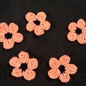 May include: Five crocheted peach-colored flowers with black centers on a dark gray background.