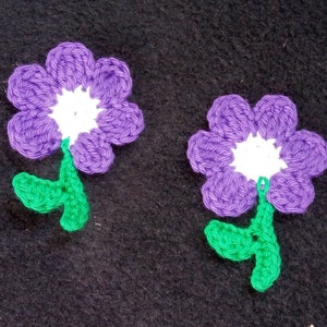 May include: Two purple crocheted flowers with white centers and green leaves. The flowers are on a black background.
