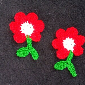 May include: Two crocheted red flowers with white centers and green leaves. The flowers are on a black background.