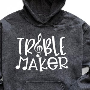 May include: A dark gray hoodie with the text "TR&BLE MAKER" in white, featuring a treble clef and musical note design. The hoodie has a hood and a front pocket. Accessories include sunglasses, sneakers, and a pair of jeans.
