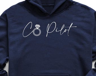 May include: Navy blue hoodie with the text "C8 Pilot" and a diamond ring graphic. The hoodie has a front pocket and a hood. The image also includes black sunglasses, black sneakers, and a pair of blue jeans.