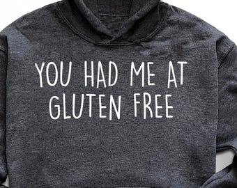 May include: Dark gray hoodie with the text "YOU HAD ME AT GLUTEN FREE" in white. The hoodie has a hood and a front pocket. The image also includes black sunglasses, black sneakers, a pair of jeans, and a small plant.