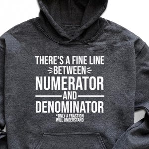 May include: Dark gray hoodie with the text "THERE'S A FINE LINE BETWEEN NUMERATOR AND DENOMINATOR. ONLY A FRACTION WILL UNDERSTAND" in white. The image also includes black sunglasses, black sneakers, and blue jeans.