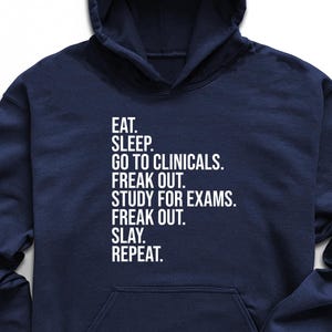 May include: Navy blue hoodie with white text: "EAT. SLEEP. GO TO CLINICALS. FREAK OUT. STUDY FOR EXAMS. FREAK OUT. SLAY. REPEAT." Black sunglasses, sneakers, and jeans are also visible. A succulent plant and a smartwatch are also in the image.