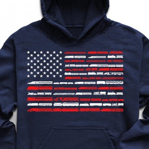 May include: Navy blue hooded sweatshirt featuring an American flag design made of train silhouettes in red, white, and blue. The sweatshirt has a front pocket and a drawstring hood. Black sneakers, sunglasses, and a watch are also in the image.