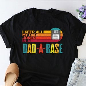 May include: Black t-shirt with a retro-style graphic that says "I keep all my dad jokes in a dad-a-base" with a floppy disk icon.