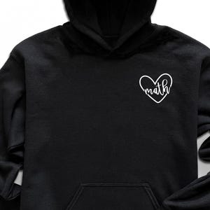 May include: Black hooded sweatshirt with a white heart graphic that says "math". The sweatshirt has a front pocket and a drawstring hood. The image also includes black sunglasses, black sneakers, a watch, and jeans.