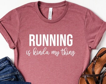 May include: A mauve t-shirt with the text "RUNNING is kinda my thing" in white lettering. The shirt is paired with blue jeans and brown sandals.