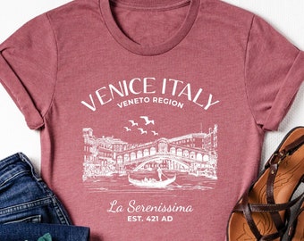 May include: A maroon t-shirt with a white graphic of Venice, Italy, and the text "Venice Italy Veneto Region La Serenissima Est. 421 AD." The shirt is paired with blue jeans, brown sandals, and a floral accessory.