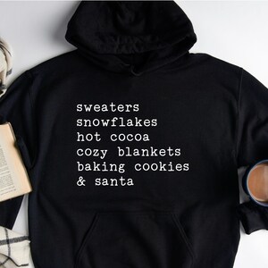 May include: Black hoodie with white text that reads "sweaters, snowflakes, hot cocoa, cozy blankets, baking cookies & santa".