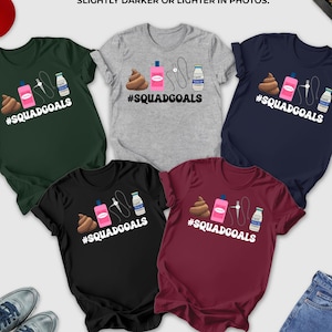 May include: A collection of t-shirts in various colors, including forest green, gray, navy, black, and maroon. Each shirt features a graphic with a poop emoji, a pink bottle, a needle, and a milk carton, with the text "#SQUADGOALS".